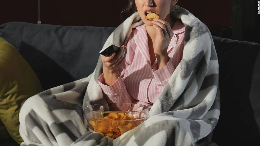 Mujer en pijama envuelta en una manta come papas fritas con control remoto en mano mientras ve televisión en el sofá.