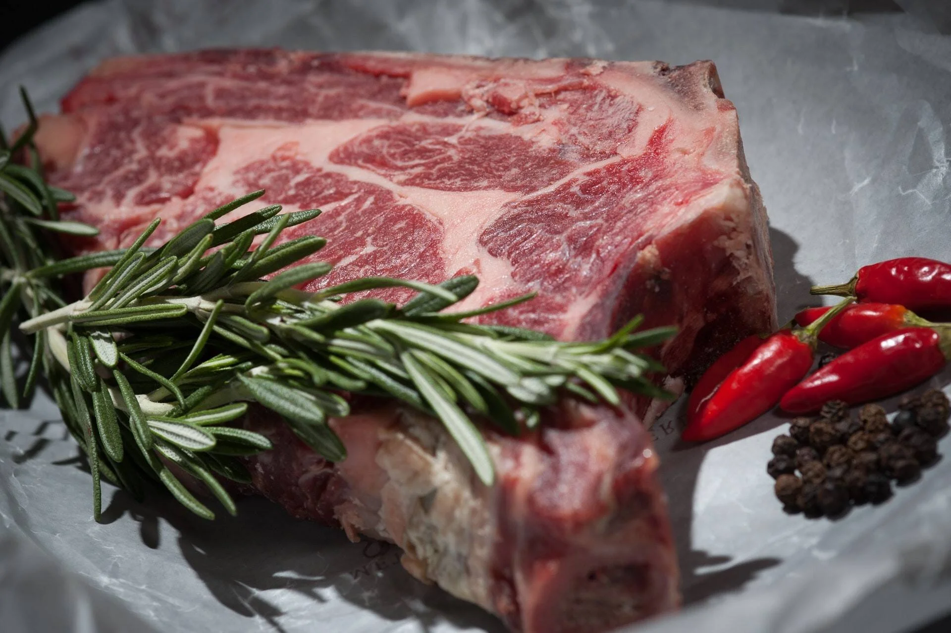 Grueso corte de carne de res cruda con marmoleado, acompañado de ramas de romero, chiles rojos y granos de pimienta negra.