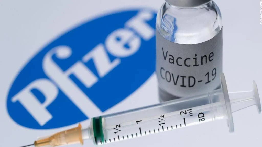 Frasco etiquetado 'Vaccine COVID-19' y una jeringa frente al logo de Pfizer.