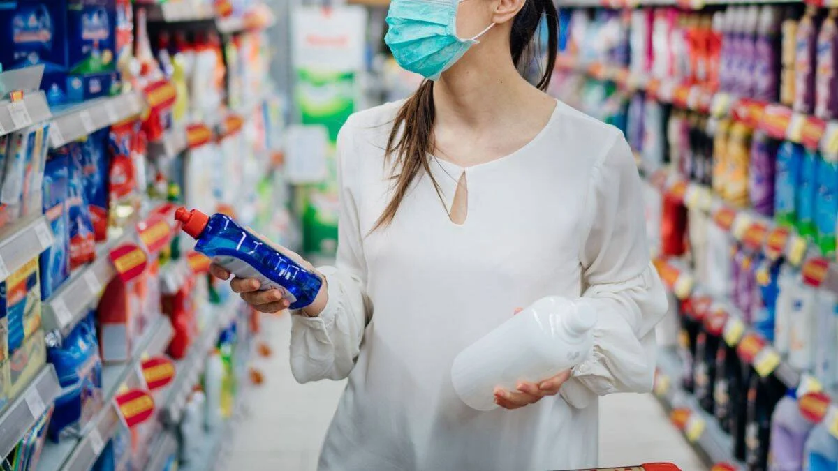 Mujer con mascarilla quirúrgica compara dos productos de limpieza en el pasillo de un supermercado.