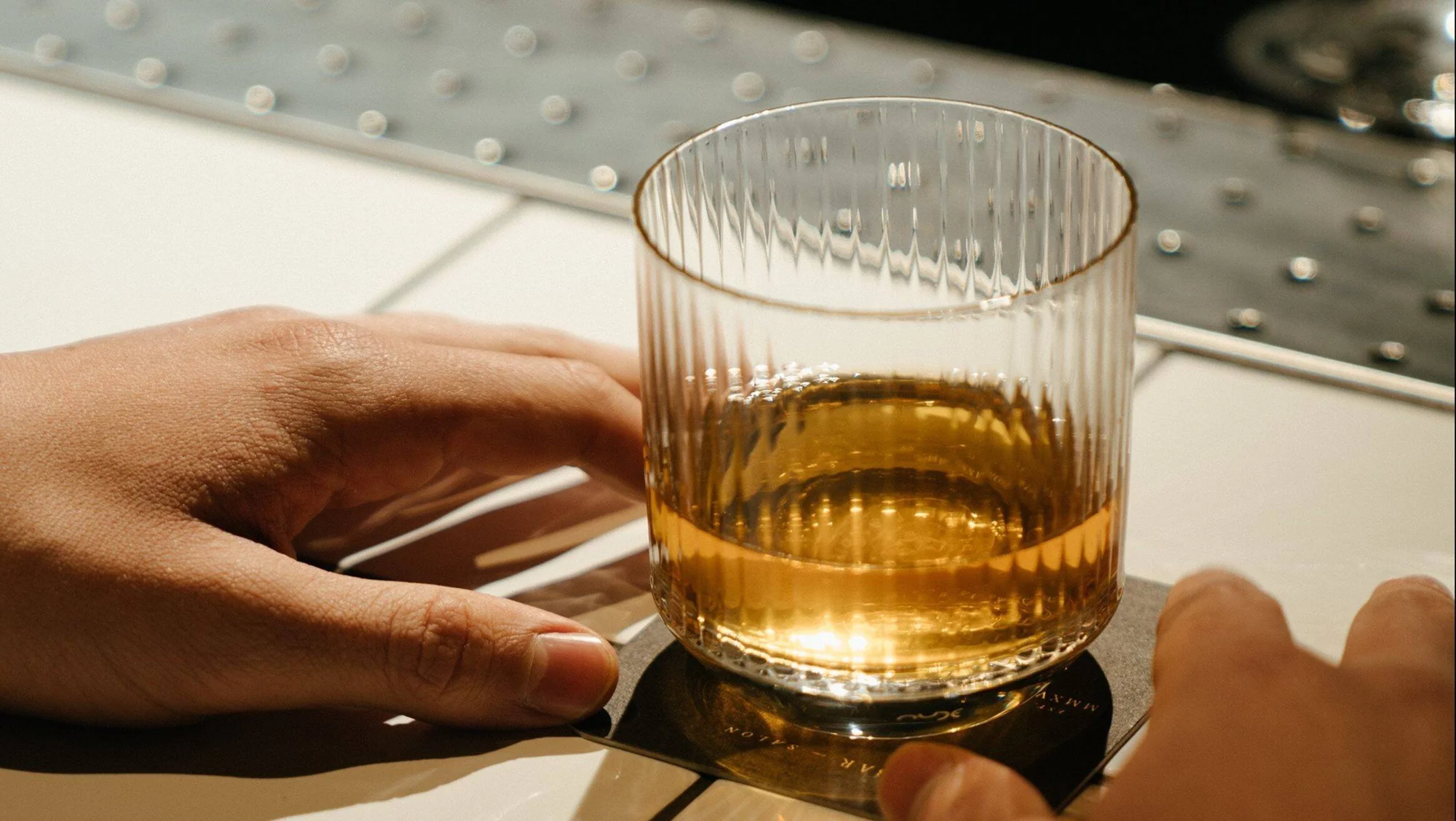 Una mano toma un vaso de cristal tallado con whisky sobre un posavasos negro, en una superficie iluminada por luz natural.
