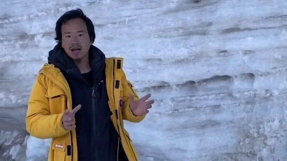 Un hombre con chaqueta amarilla habla gesticulando frente a una pared de hielo o glaciar.