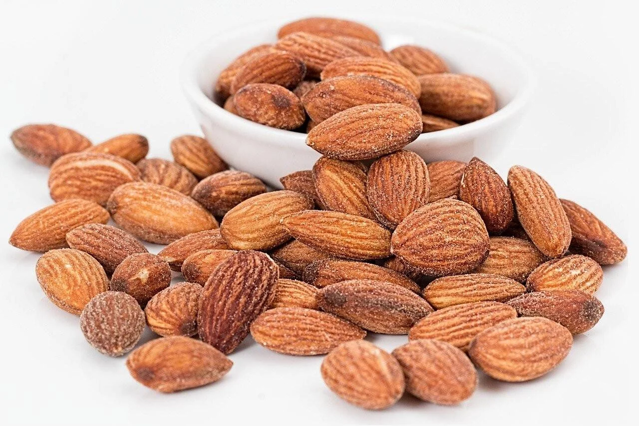 Puñado de almendras tostadas esparcidas sobre superficie blanca junto a un cuenco lleno del mismo fruto seco.