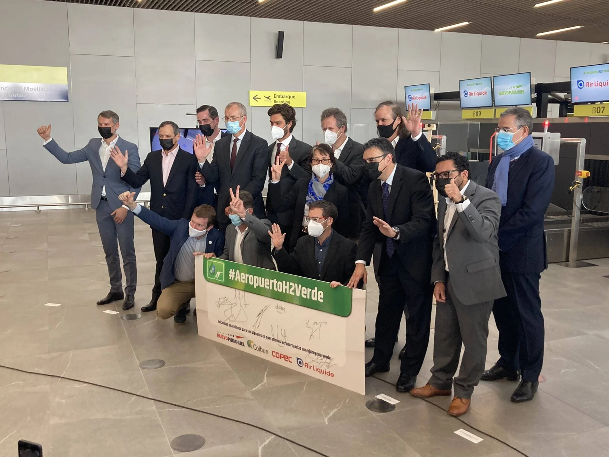 Grupo de ejecutivos con mascarilla posa en el aeropuerto sosteniendo un cartel con el hashtag #AeropuertoH2Verde, alianza entre Nuevo Pudahuel, Colbún, Copec y Air Liquide.
