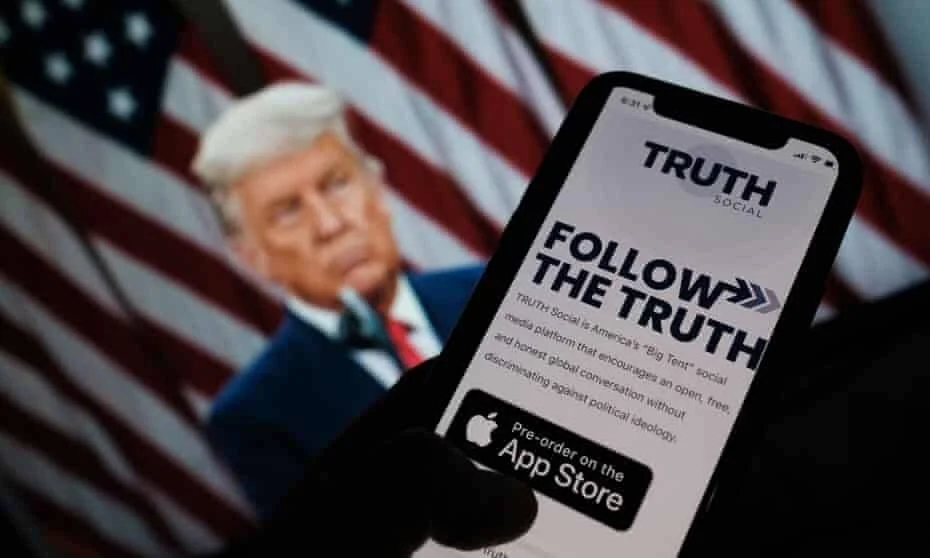 Mano sostiene un teléfono con la app Truth Social en pantalla, con la imagen de Trump y una bandera estadounidense desenfocadas al fondo.