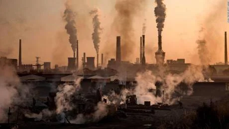 Complejo industrial con múltiples chimeneas expulsando grandes columnas de humo en un cielo anaranjado oscuro.