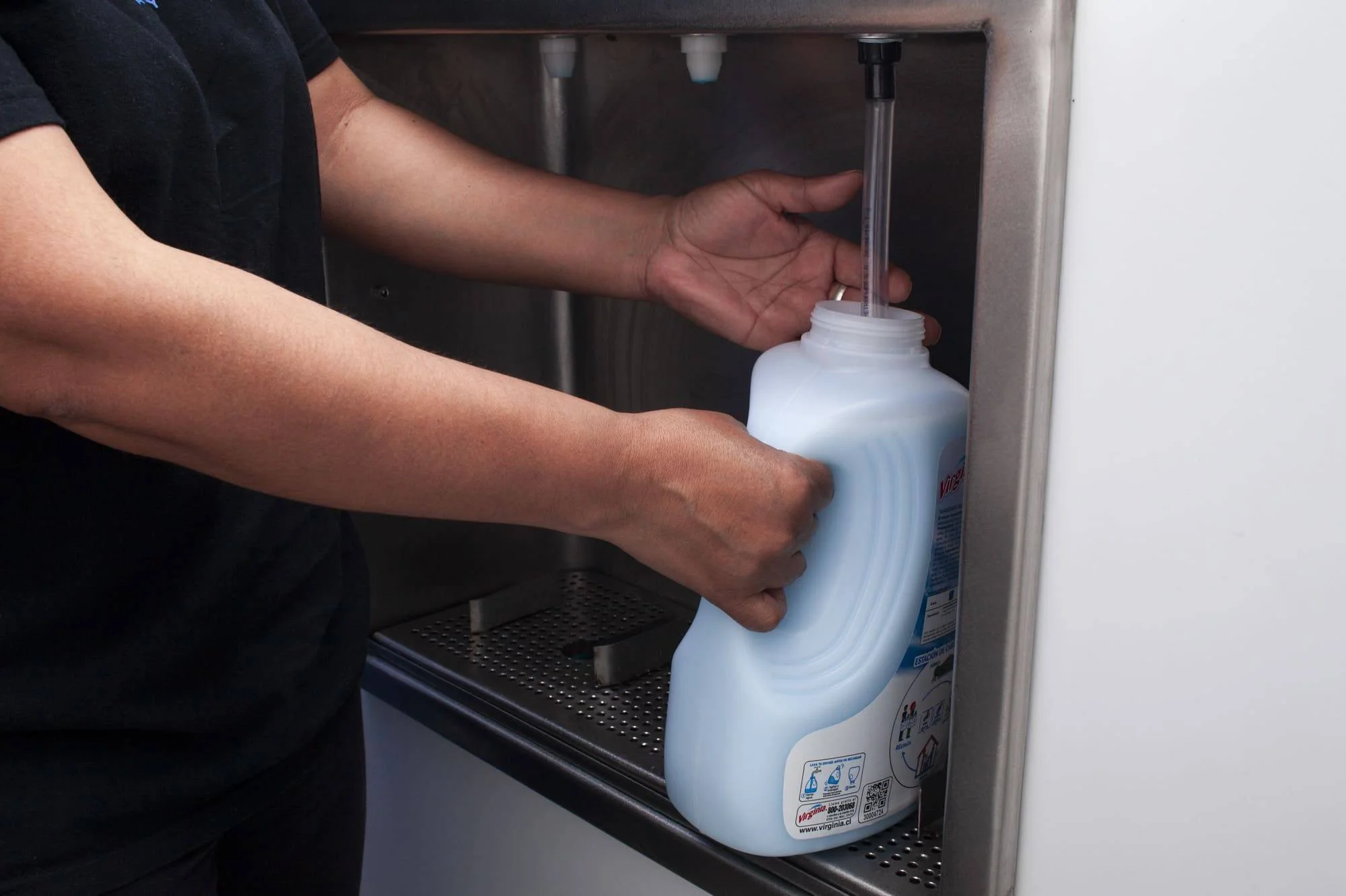 Manos sosteniendo un envase plástico de detergente líquido mientras lo recargan en una estación dispensadora de acero inoxidable.