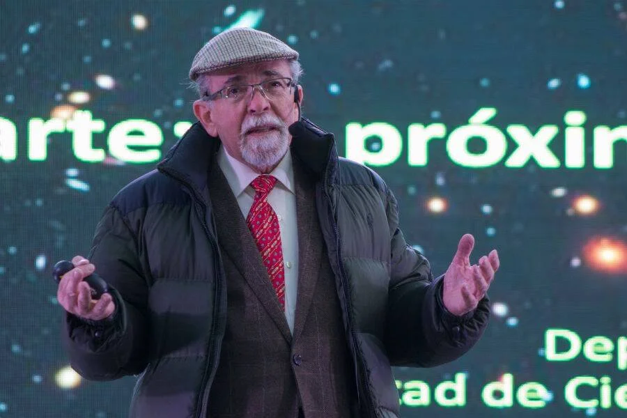Hombre mayor con gorra y corbata roja gesticula durante una presentación frente a una pantalla con texto en verde.