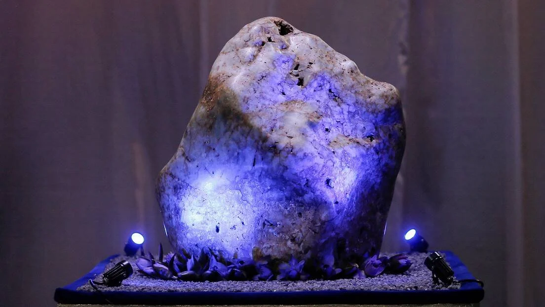 Enorme roca de zafiro en bruto iluminada con focos azules sobre una base decorativa con pétalos y arena.