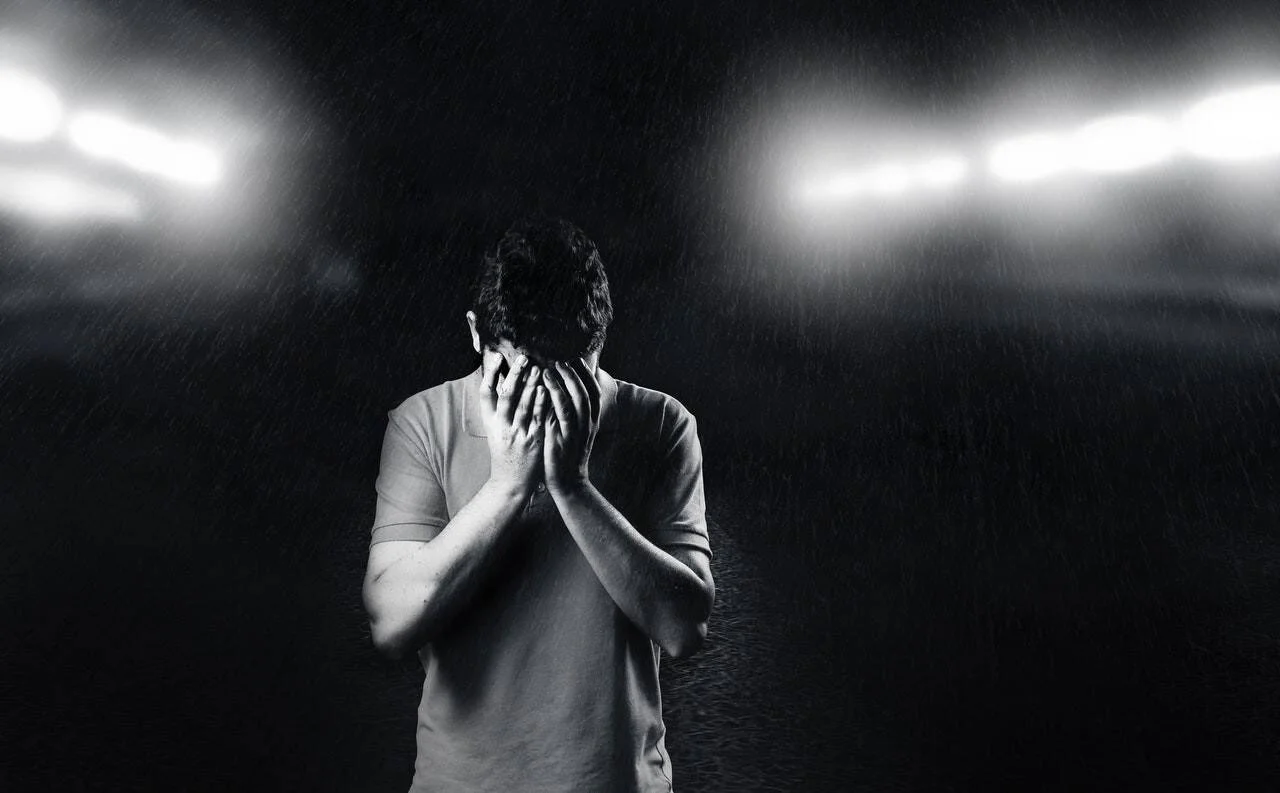 Hombre con el rostro cubierto entre sus manos, bajo la lluvia y la luz de focos de estadio, en blanco y negro.