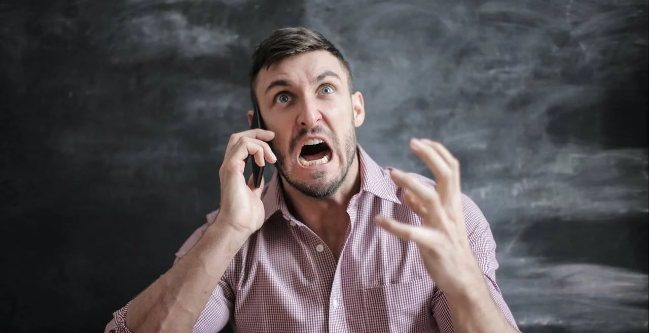 Hombre con expresión de enojo grita mientras habla por teléfono celular y gesticula con la mano.