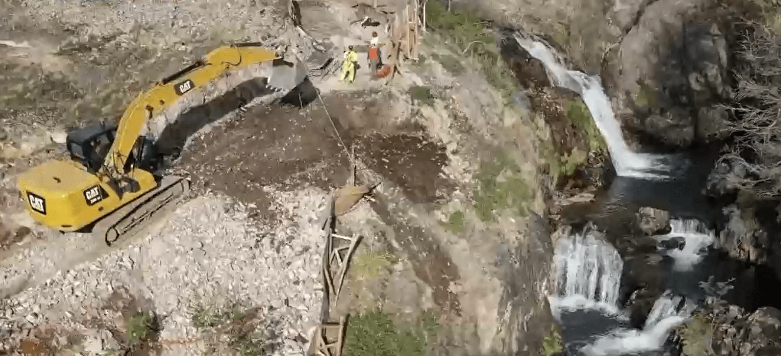 Excavadora CAT amarilla opera en terreno rocoso junto a una cascada, con trabajadores supervisando las obras desde altura.