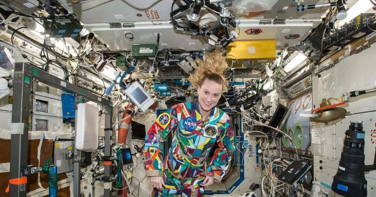 Astronauta de la NASA con traje multicolor flota sonriendo en el interior de la Estación Espacial Internacional, rodeada de equipos y cables.