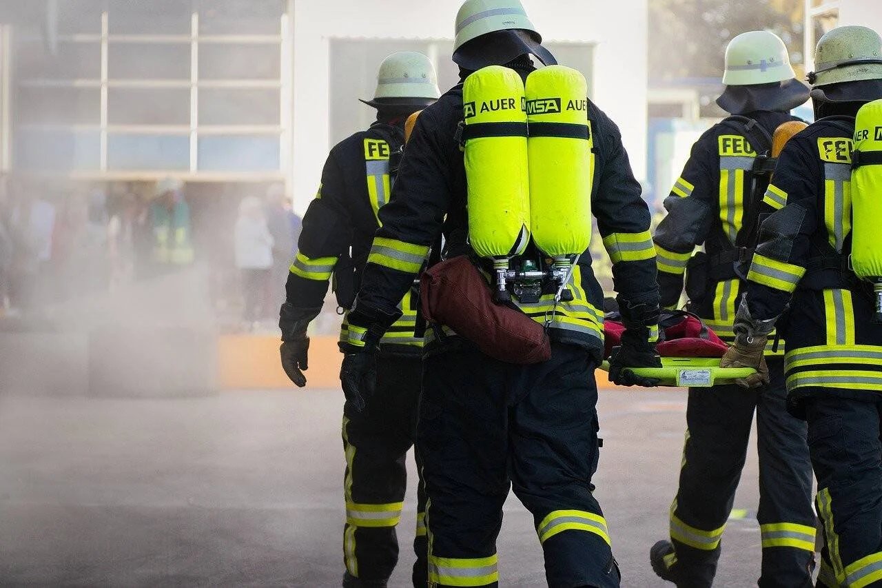 Varios bomberos equipados con trajes ignífugos y botellas de aire comprimido transportan una camilla entre humo durante una intervención de emergencia.