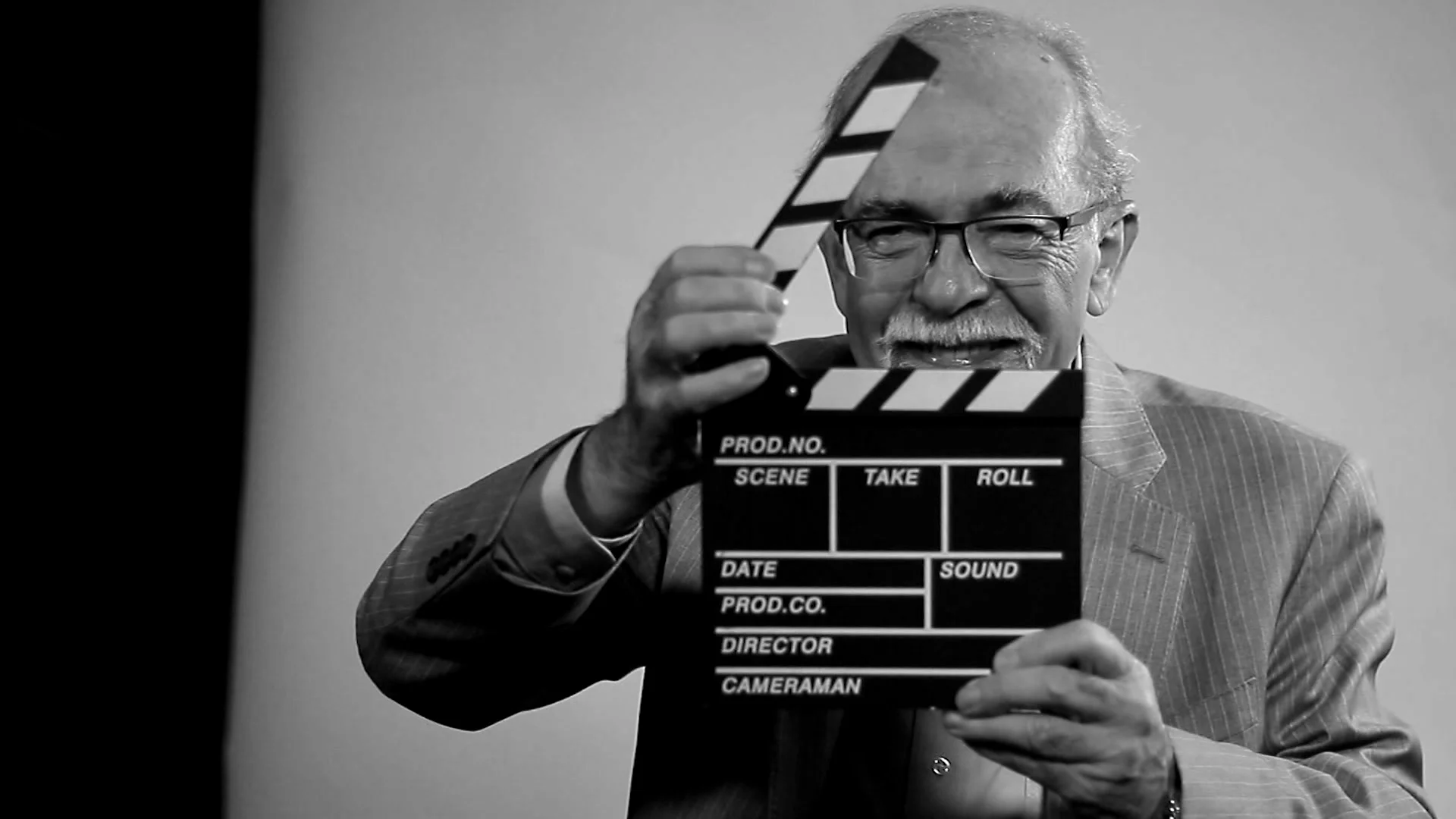 Hombre mayor con lentes y traje sostiene una claqueta de filmación frente a su rostro, en blanco y negro.