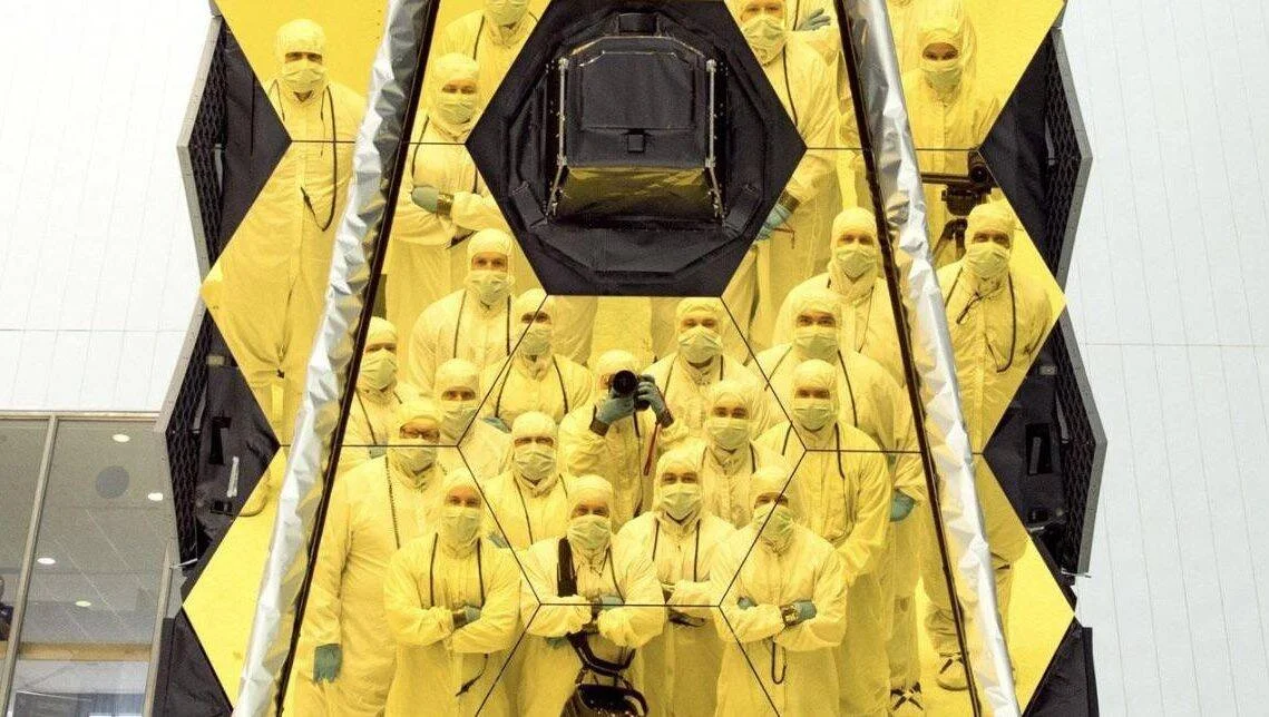 Grupo de ingenieros con trajes de sala limpia posando frente al espejo primario hexagonal dorado de un telescopio espacial.