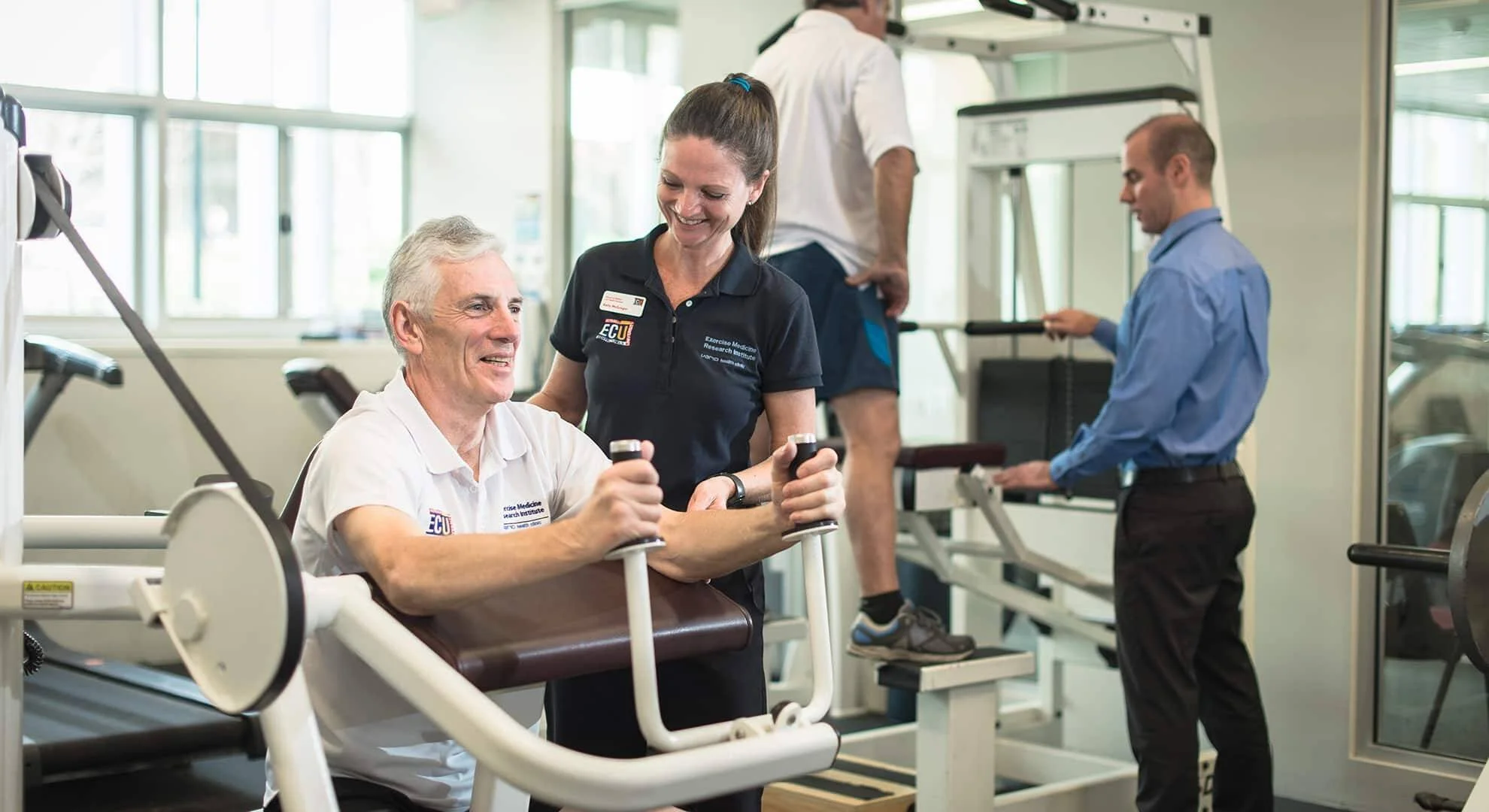 Profesional del Exercise Medicine Research Institute asiste a un hombre mayor en una máquina de ejercicio en un gimnasio clínico.