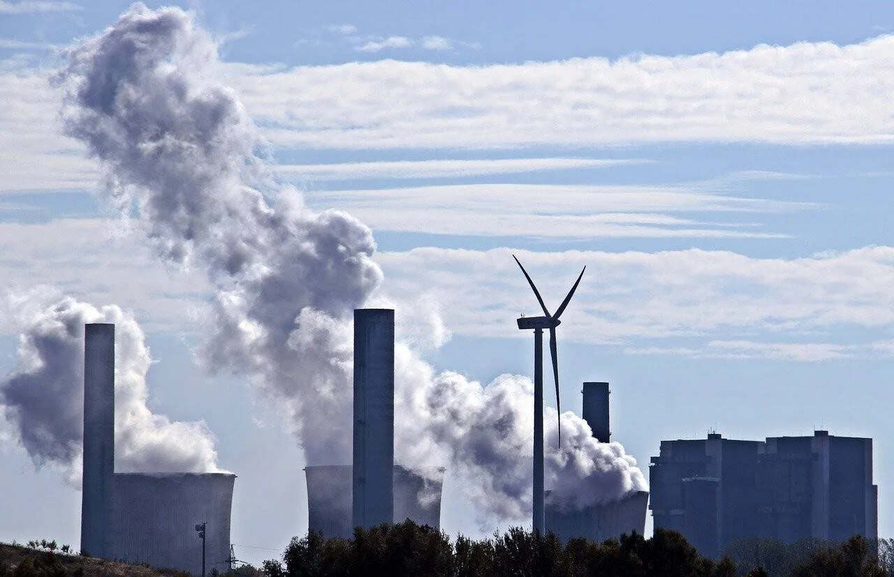 Aerogenerador junto a una central térmica que emite grandes columnas de humo, simbolizando la tensión entre energías fósiles y renovables.