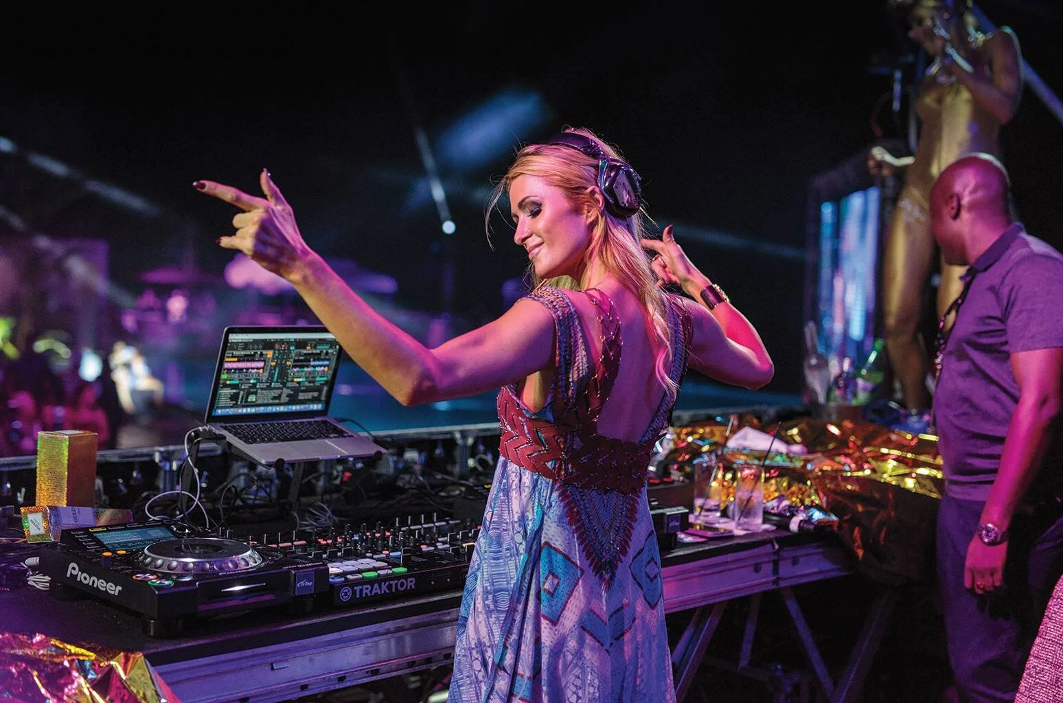 DJ rubia con auriculares señala al público desde su cabina con equipos Pioneer y Traktor en una discoteca iluminada.