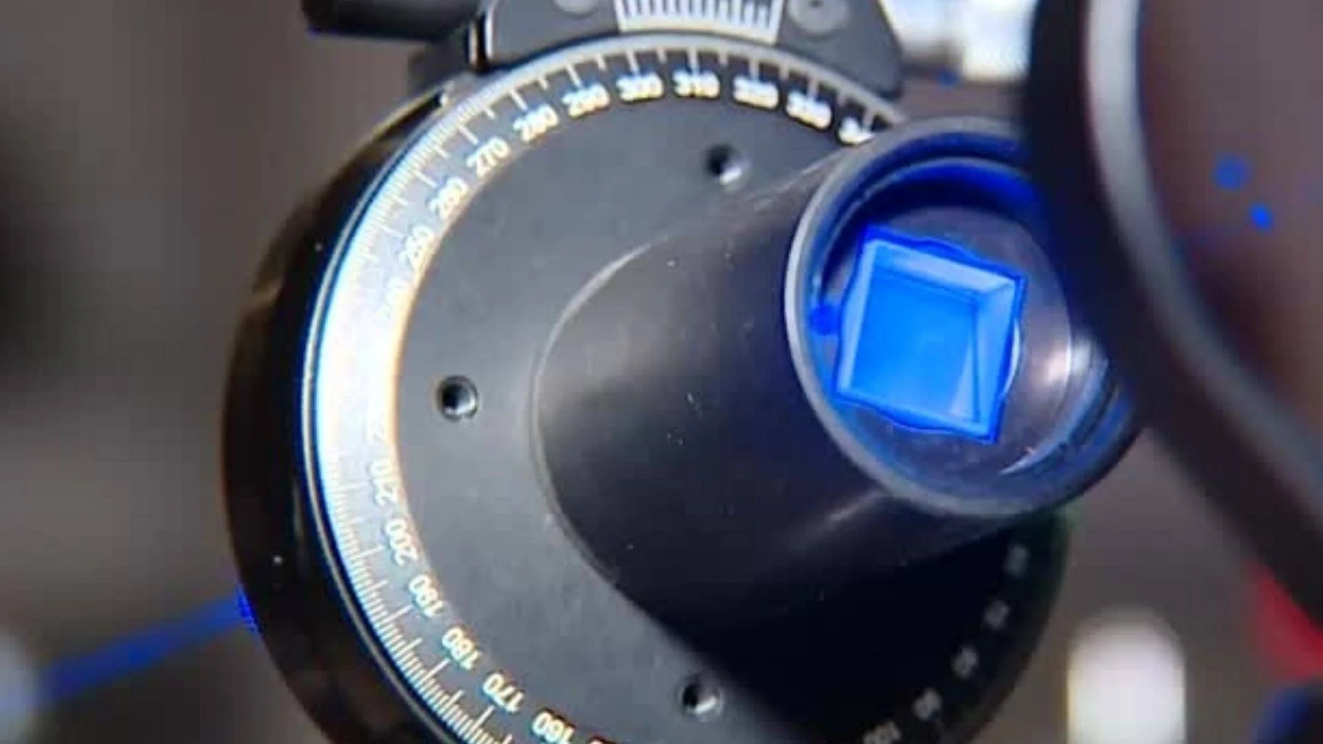 Primer plano de un instrumento de precisión con escala graduada en grados y un elemento óptico iluminado en azul.