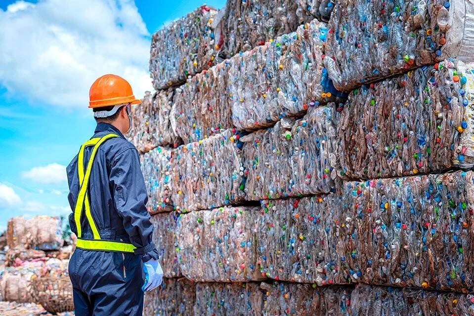 Operario con casco y chaleco de seguridad observa grandes fardos de botellas plásticas compactadas en planta de reciclaje.
