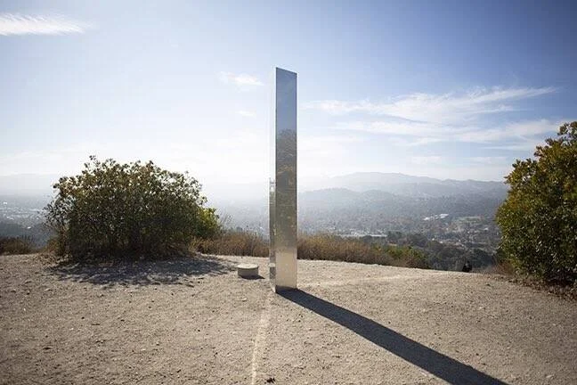 Columna metálica espejada de gran altura instalada en una colina árida con una ciudad y montañas al fondo.