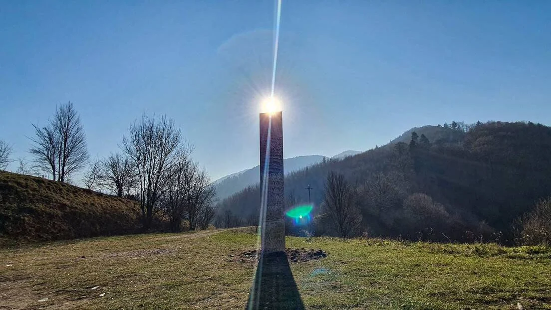 Monolito metálico brillante en campo abierto con montañas al fondo y sol reflejándose en su cúspide.