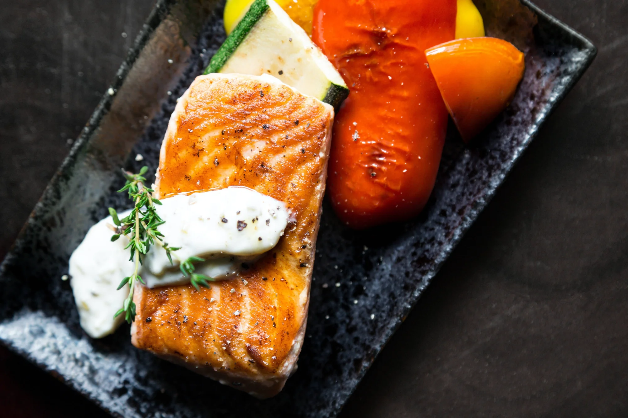 Filete de salmón dorado con salsa cremosa y tomillo, acompañado de tomates y calabacín asados en plato negro.