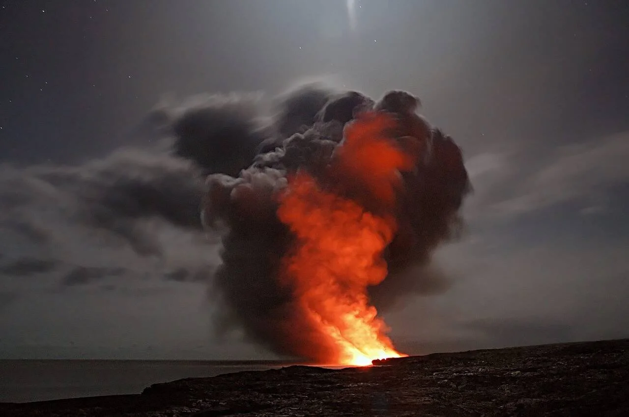 Lava incandescente genera una gigantesca columna de humo naranja y negro sobre la costa rocosa durante la noche.