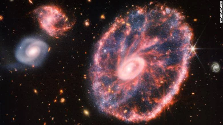 La Galaxia Rueda de Carro y dos galaxias compañeras fotografiadas en infrarrojo, mostrando anillos rosados y estructuras en el espacio profundo.