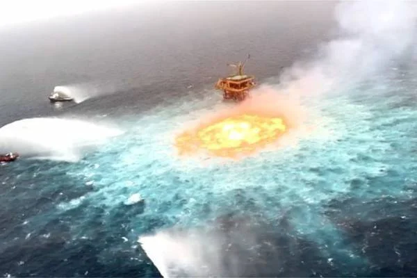 Gran llamarada ardiendo sobre la superficie del océano junto a una plataforma petrolera, mientras embarcaciones combaten el fuego con agua.