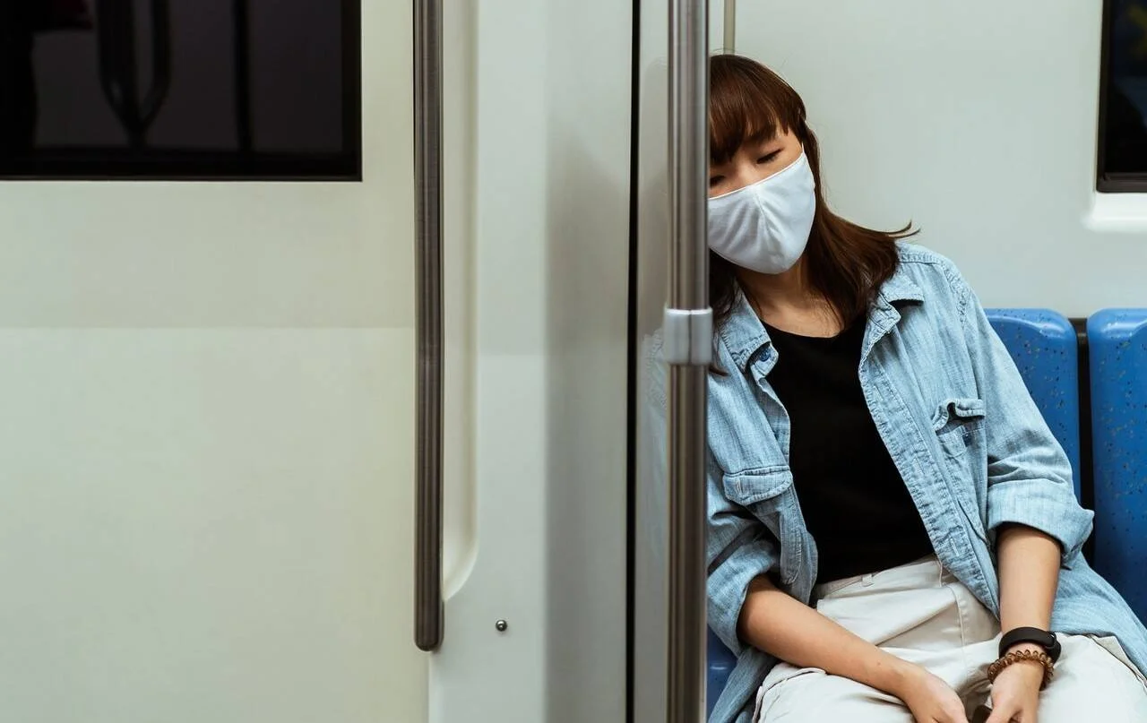 Mujer joven con mascarilla blanca y ojos cerrados, dormida en un asiento de transporte público.