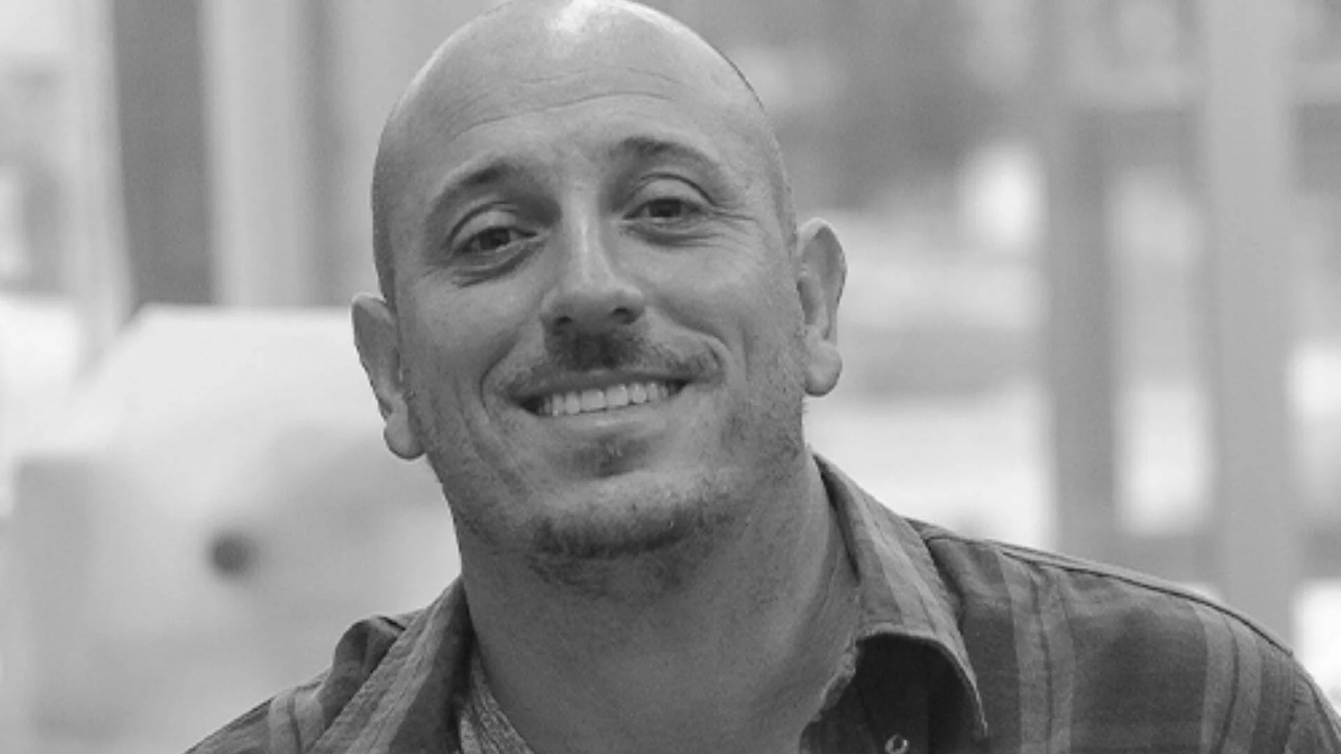 Hombre de cabeza rapada y barba incipiente sonríe a cámara en fotografía en blanco y negro.