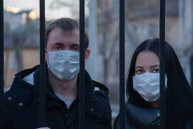 Un hombre y una mujer con mascarillas quirúrgicas miran a través de una reja metálica en un espacio exterior.