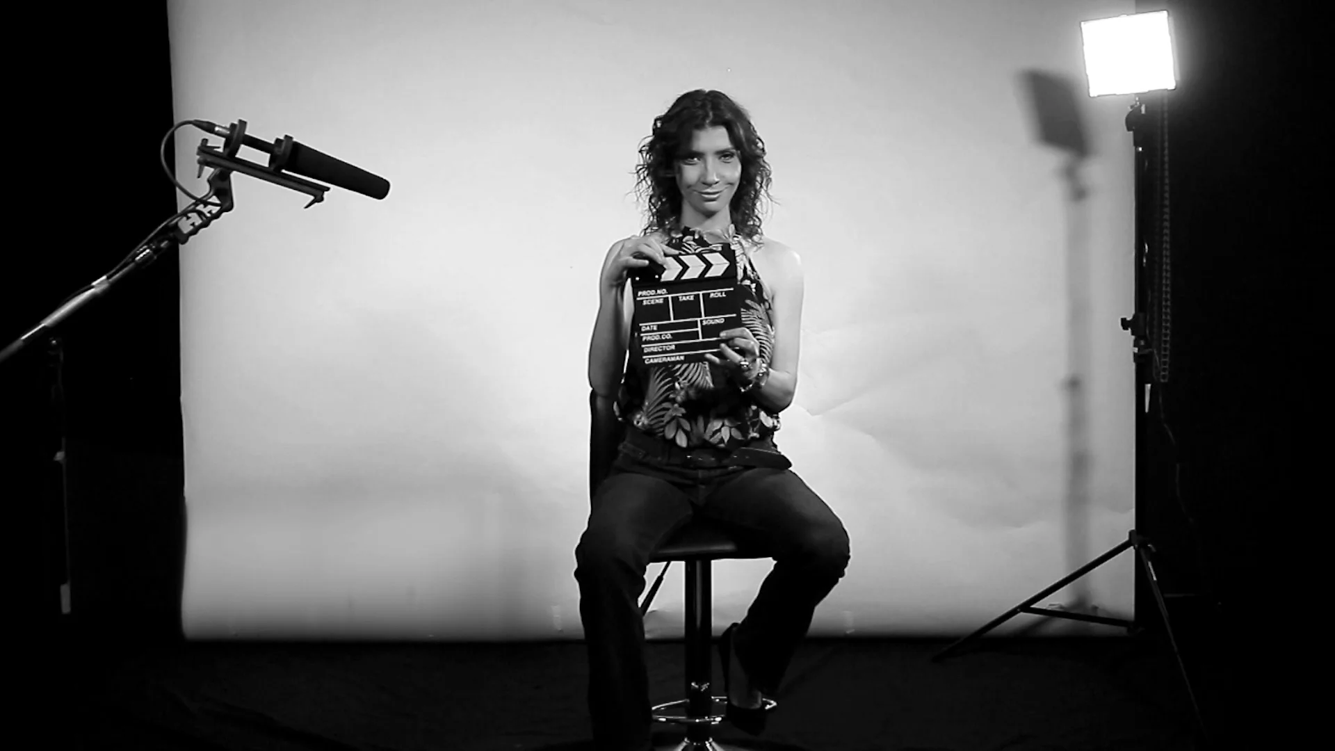 Mujer sentada en taburete sosteniendo una claqueta de cine en un estudio con micrófono boom y luz de producción.