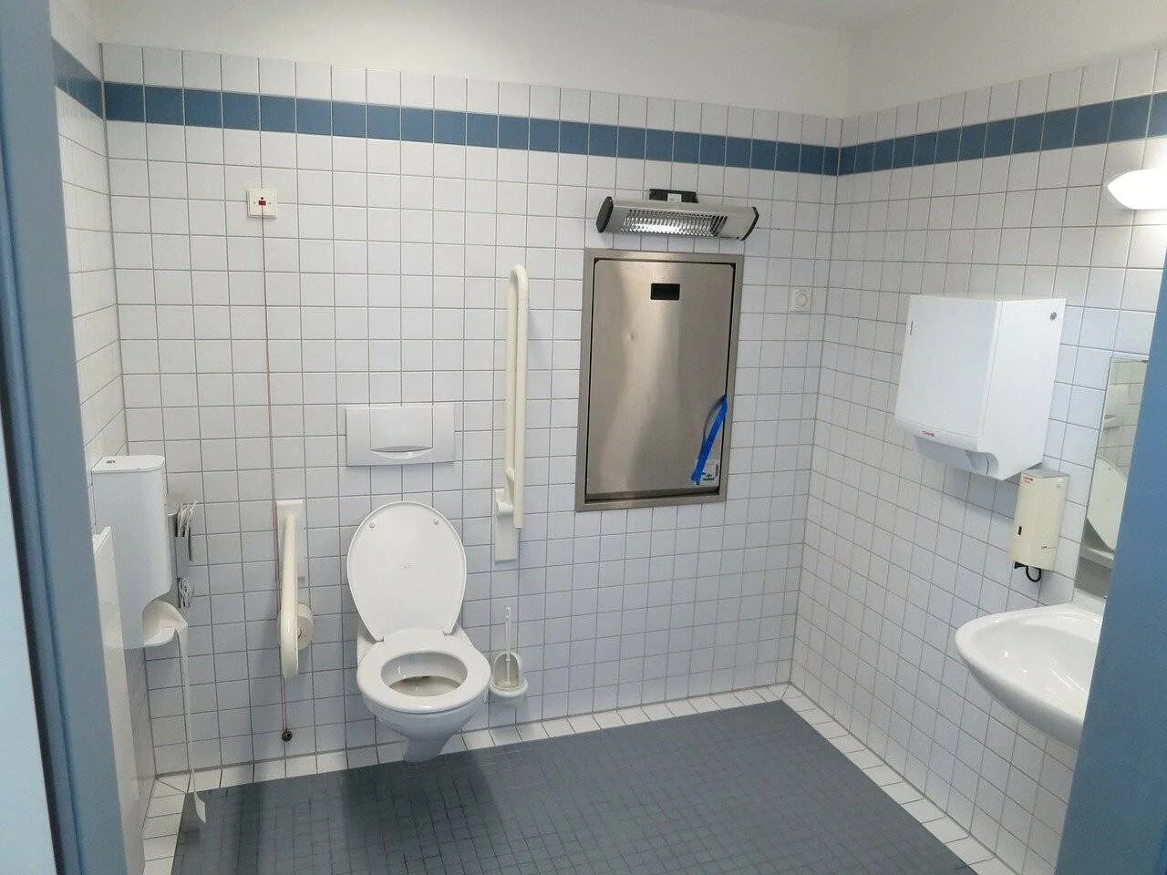 Baño adaptado para personas con discapacidad con inodoro, barras de apoyo, dispensadores de papel y cambiador de bebés empotrado.