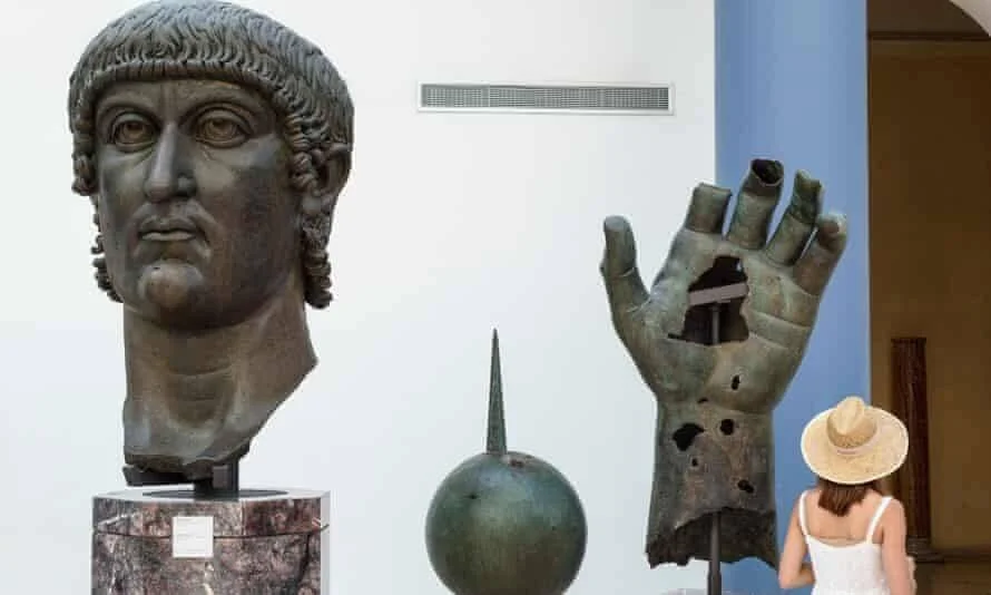 Una visitante con sombrero observa los fragmentos de bronce de un coloso romano, incluyendo una cabeza y una mano gigantes, expuestos en un museo.