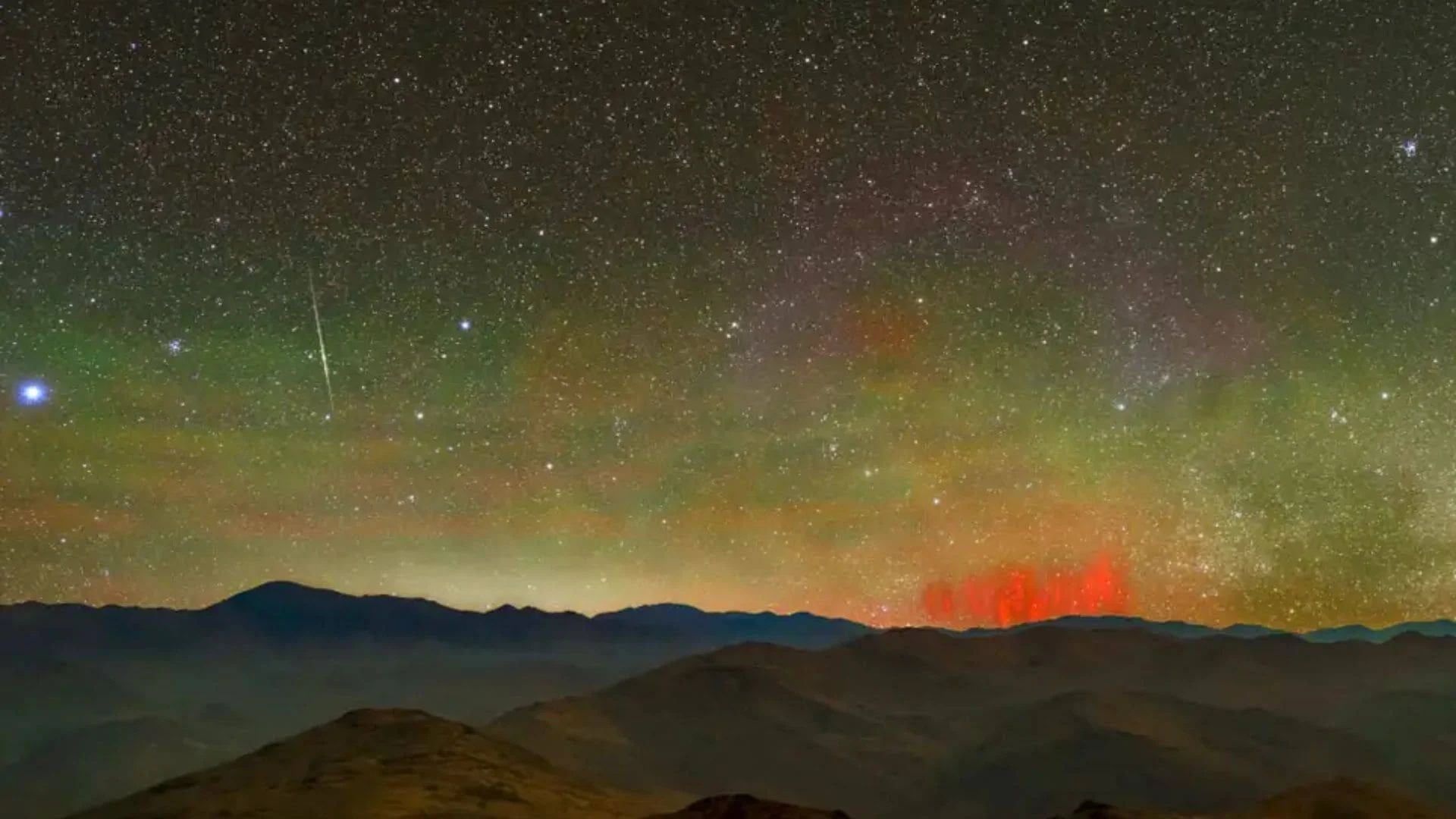 Cielo nocturno lleno de estrellas con un meteoro, auroras verdes y columnas rojas de luz sobre una cordillera oscura.