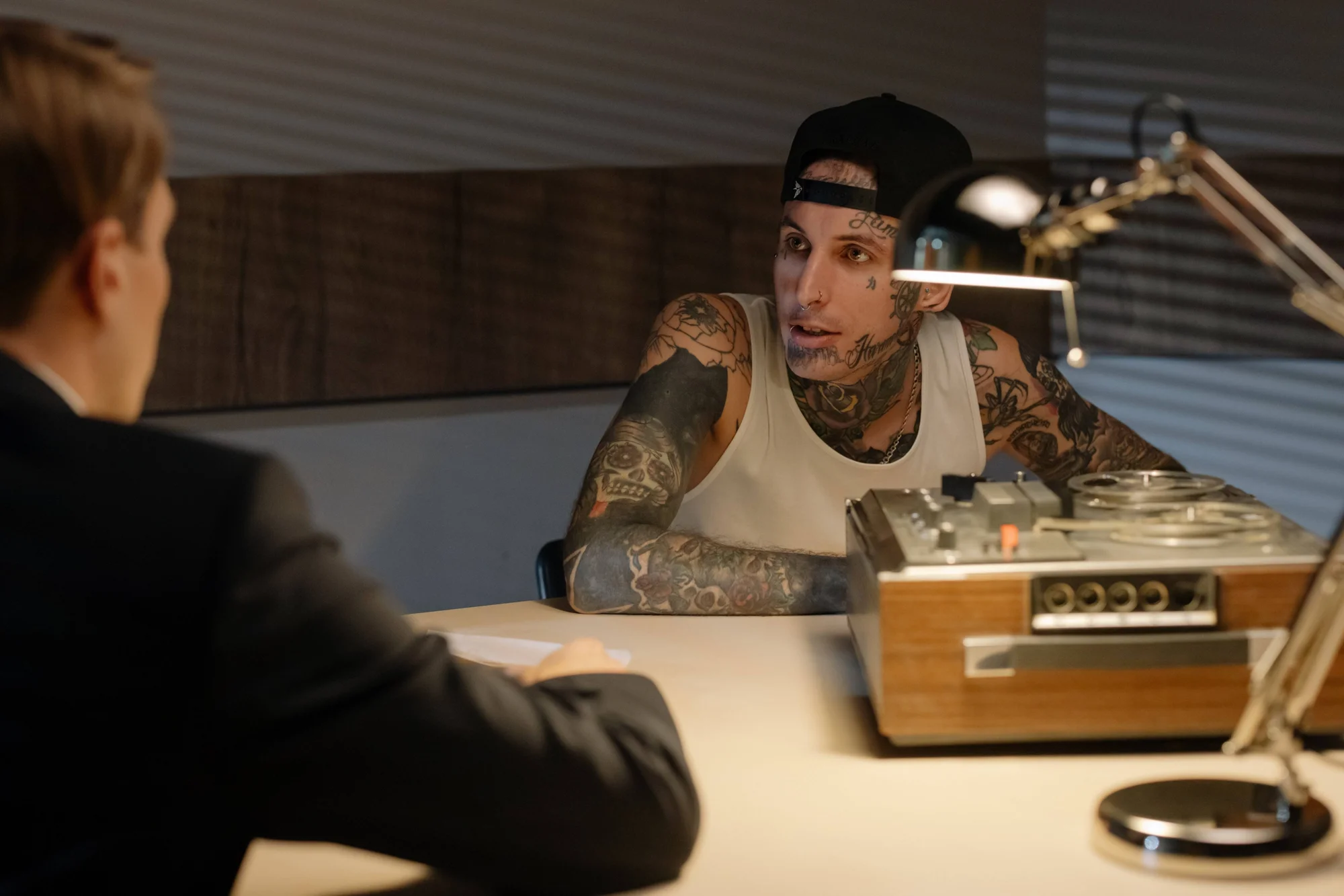 Hombre con tatuajes en cara y brazos, gorra negra y musculosa blanca, inclinado sobre una mesa frente a otra persona en lo que parece un interrogatorio.