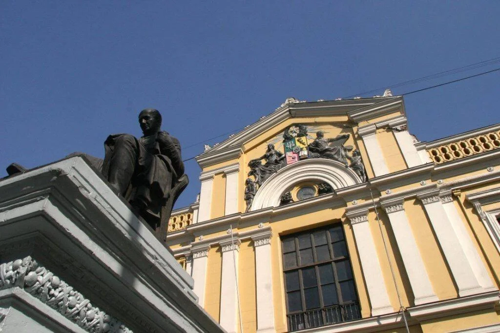 Estatua de bronce sobre pedestal ornamentado junto a la fachada neoclásica amarilla de un edificio con escudo de armas y figuras alegóricas.