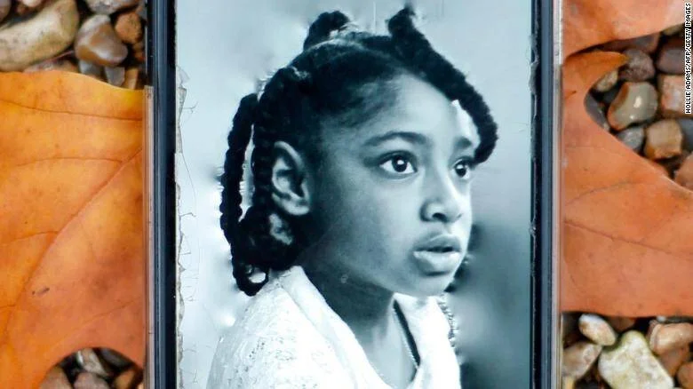Foto en blanco y negro de una niña colocada entre piedras y hojas otoñales, posiblemente en un memorial.