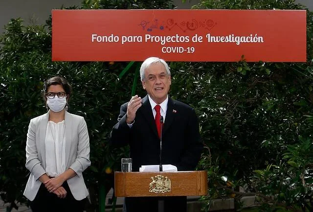 Hombre en podio con bandera chilena habla ante cartel del Fondo para Proyectos de Investigación COVID-19, junto a mujer con mascarilla.