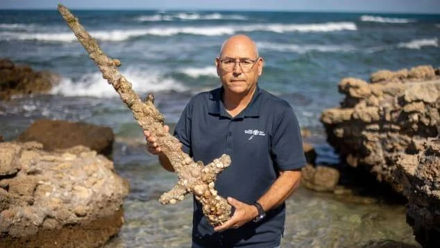 Hombre de mediana edad muestra una espada antigua cubierta de incrustaciones marinas frente al mar en una costa rocosa.