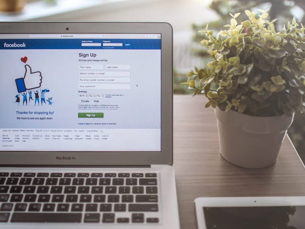 MacBook Air con la página de registro de Facebook abierta, junto a una planta y una tablet sobre un escritorio.