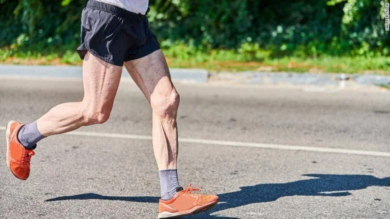 Piernas musculosas de una persona mayor corriendo en una calle de asfalto con zapatillas deportivas naranjas.