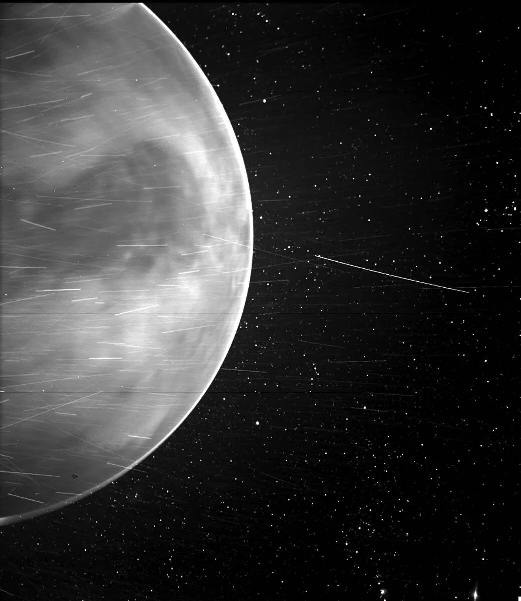 Vista en blanco y negro de Venus iluminado contra el espacio estrellado, con rastros de partículas captados por una sonda espacial.