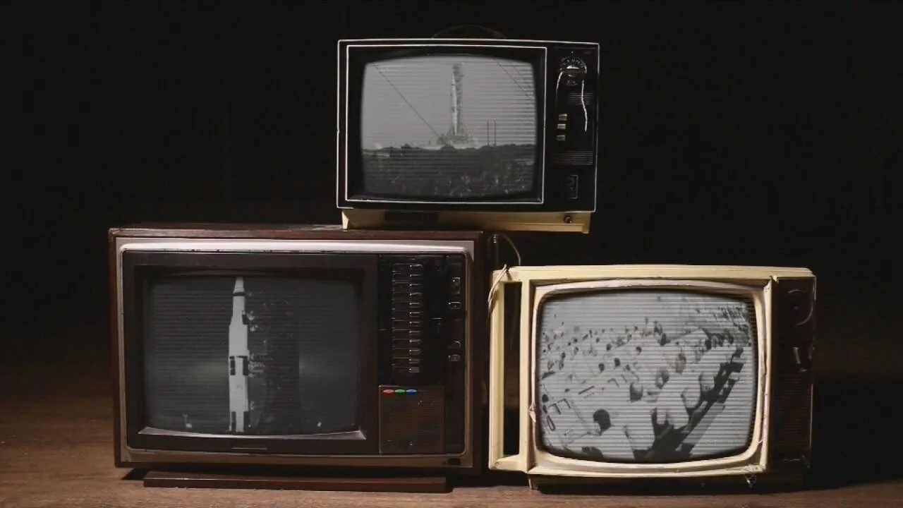 Tres televisores vintage en blanco y negro apilados muestran imágenes de un cohete espacial y una multitud.