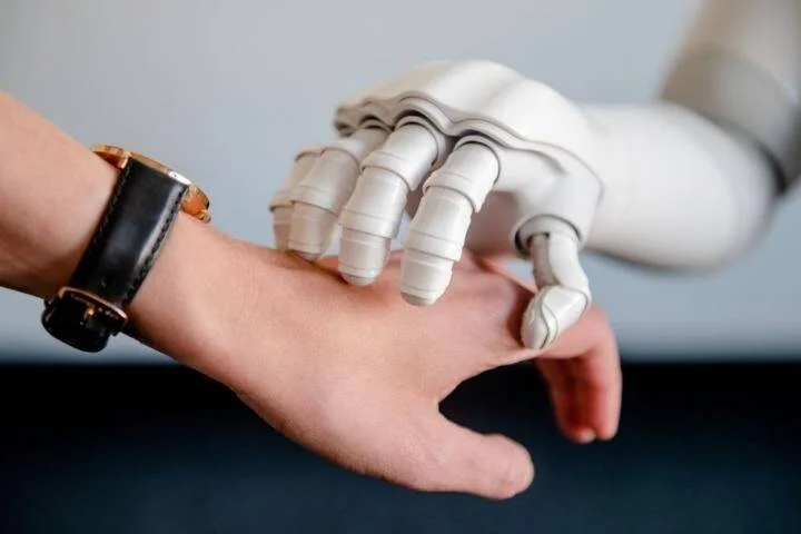 Una mano robótica blanca toca suavemente la mano de una persona que lleva un reloj de pulsera, simbolizando la interacción humano-máquina.