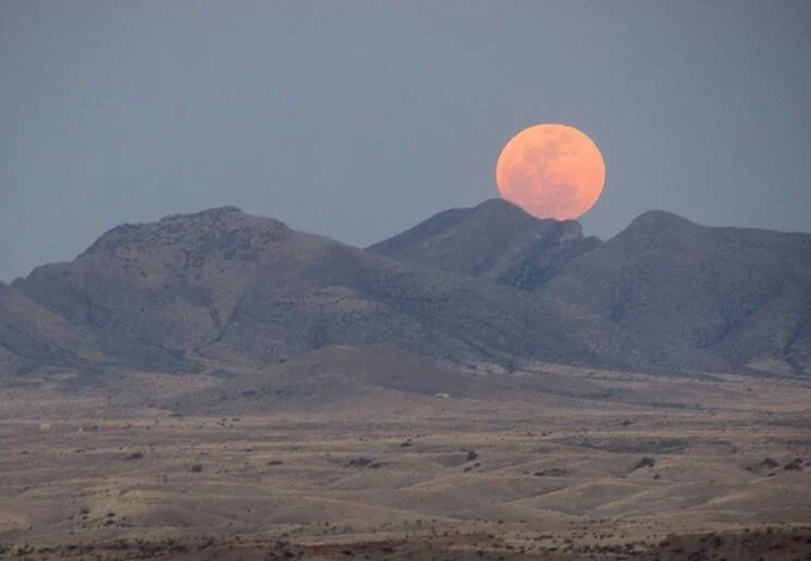 Luna llena de color naranja intenso asomando sobre una cadena montañosa en un paisaje desértico árido.