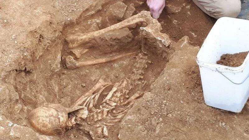 Arqueólogo excavando restos óseos humanos completos en una fosa de tierra rojiza durante una excavación arqueológica.